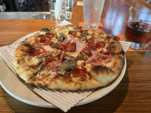 Thin crust pizza with bacon, pepperoni, and sausage on a plate. Cocktail in the top right corner of the picture.