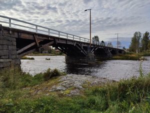 A rustic wooden bridge with stone pillars spans a flowing river.