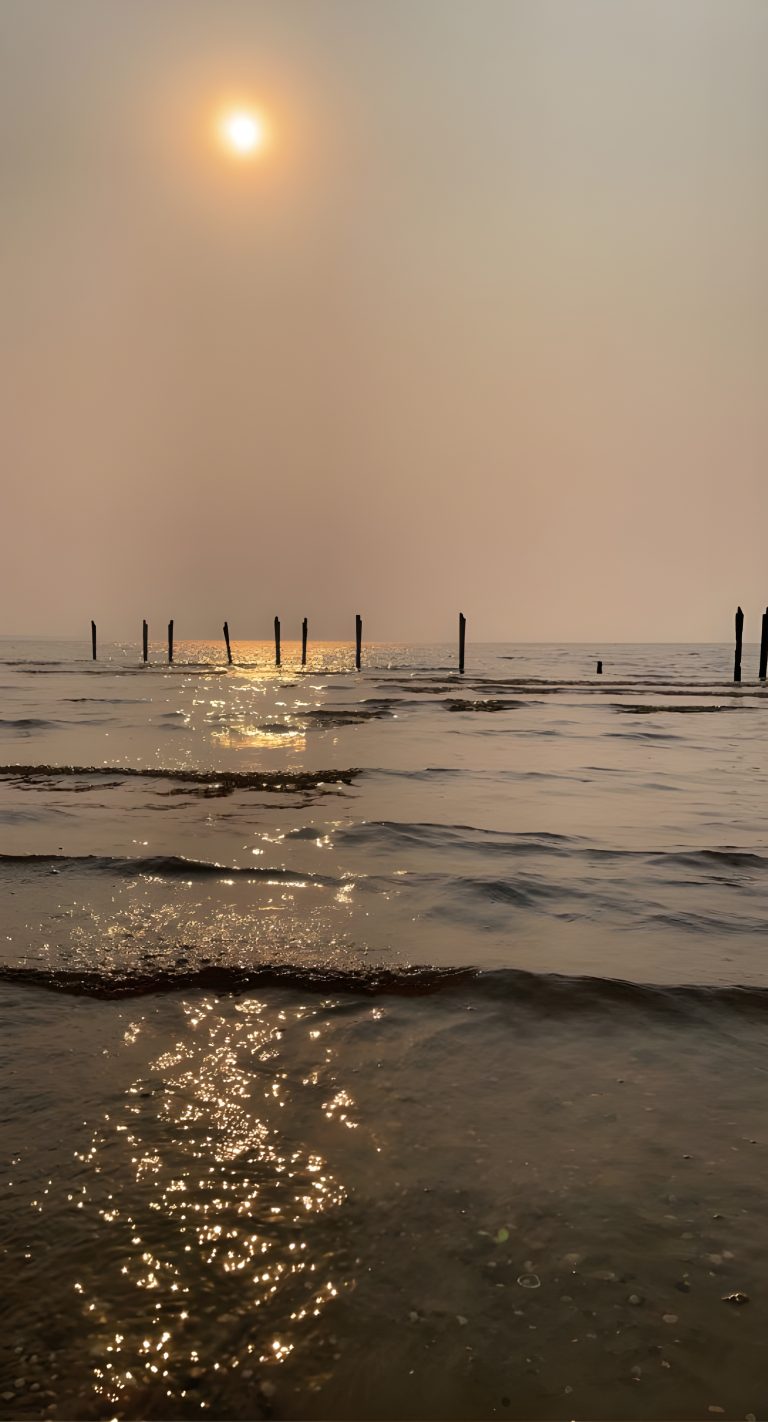 A tranquil seascape during sunset with the sun partially obscured by haze, casting a soft golden glow over the water. Old wooden posts stand scattered in the sea, and the gentle waves shimmer with reflections of sunlight. “Lago Ypacarai” in paraguay.