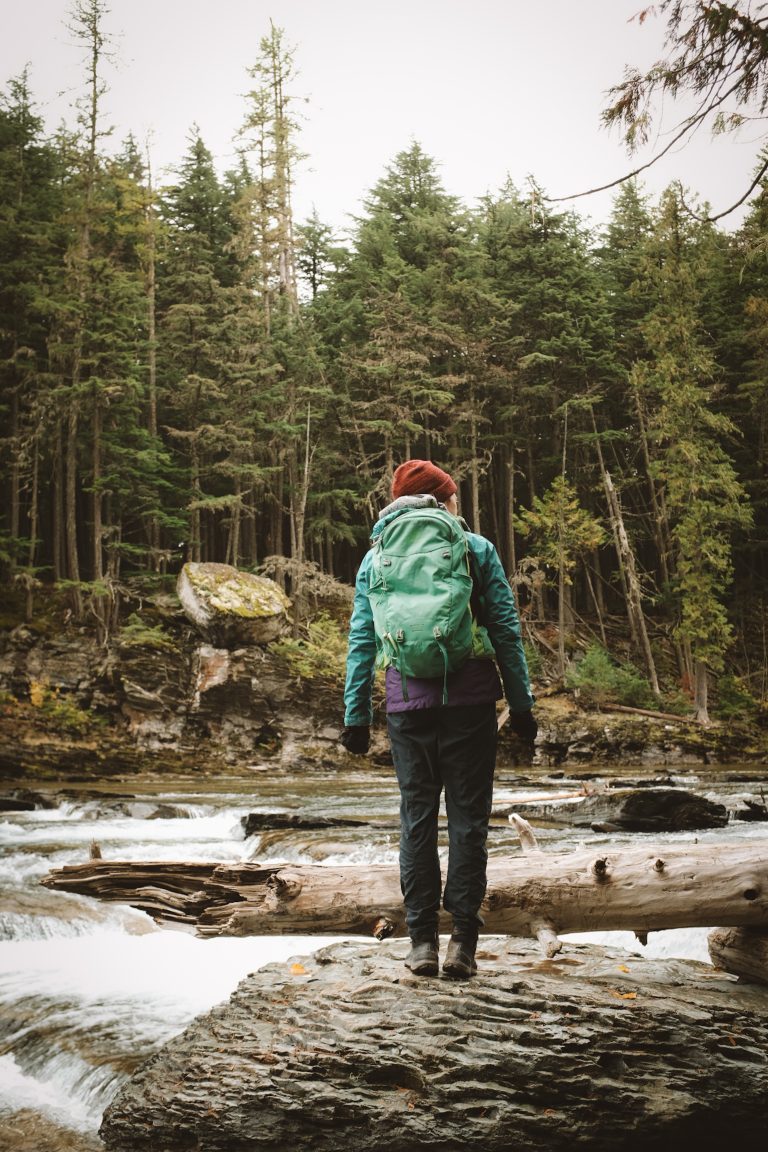 Person standing in front of a river surrounded by forest trees in winter gear, looking out on the water.