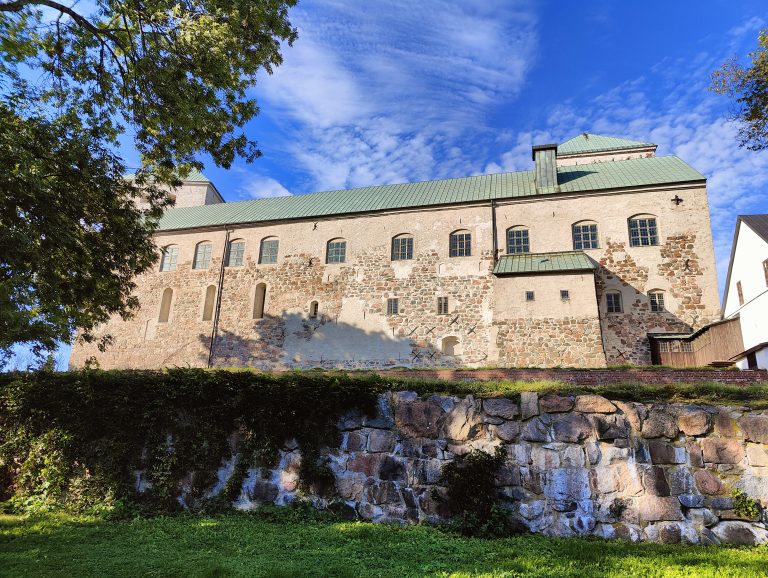 The image displays a close-up view of Turku Castle’s side facade, highlighting its textured stone walls and sturdy windows, atop a stone foundation with ivy. The castle, bathed in sunlight, contrasts with the partly cloudy sky.