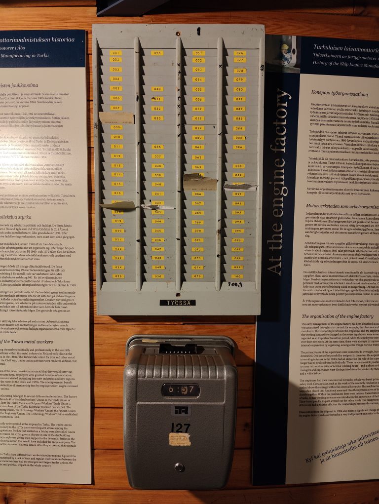 The image shows an old-fashioned punch clock with a timestamp of 6:57 and a display board with numerous in/out slots for time cards, indicating it’s part of a historical exhibit, likely on industrial labor.