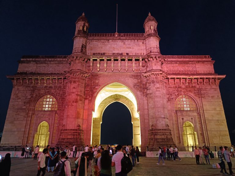 Gateway of india, mumbai during the night light, there are many tourists standing in front of the gate. On the front there is very dimm lighting and inside the gate there is very bright lighting.