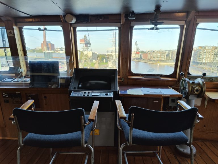 A ship’s bridge with two empty chairs facing a wheel and navigational equipment, overlooking a waterway with industrial structures in the distance.