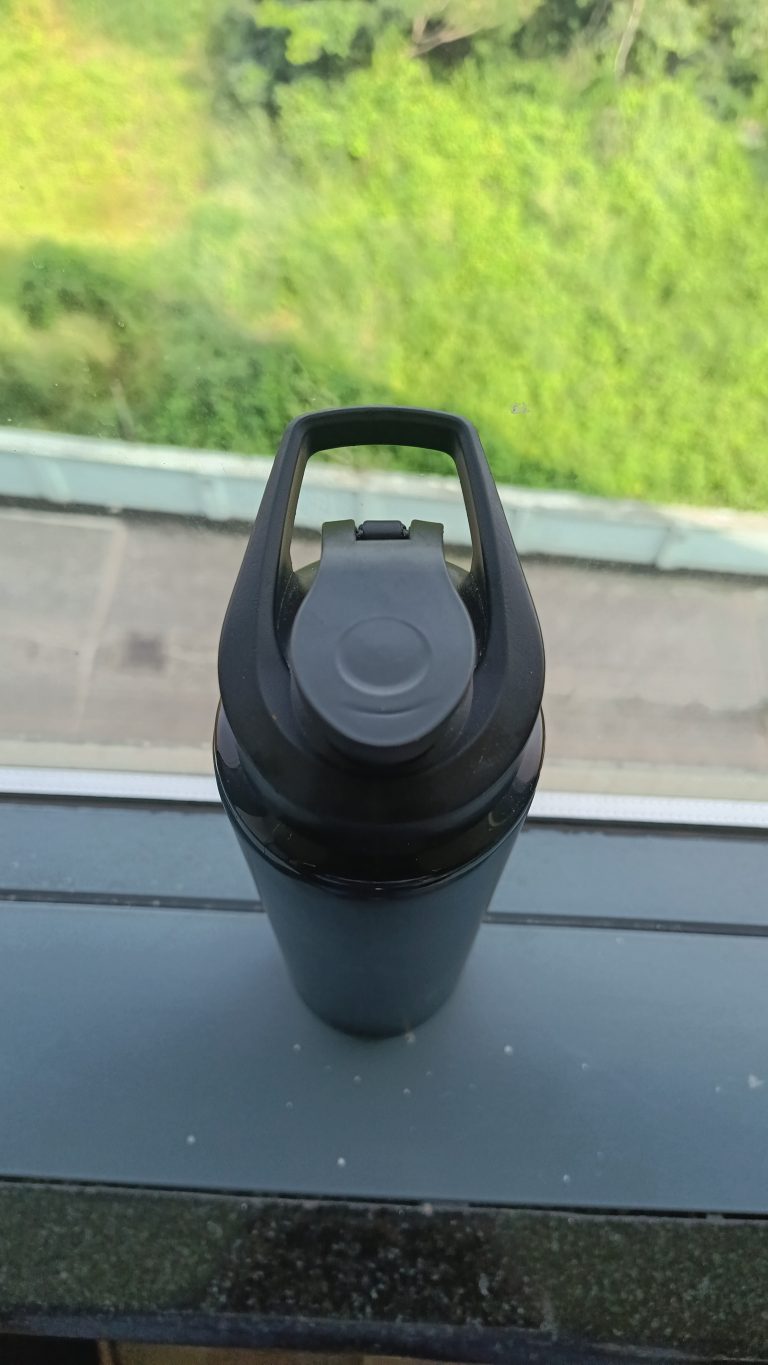 A black water bottle placed near the glass window and a green scenery behind it.