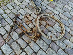 A severely rusted and deteriorated bicycle frame and wheel lie on a cobblestone path. 