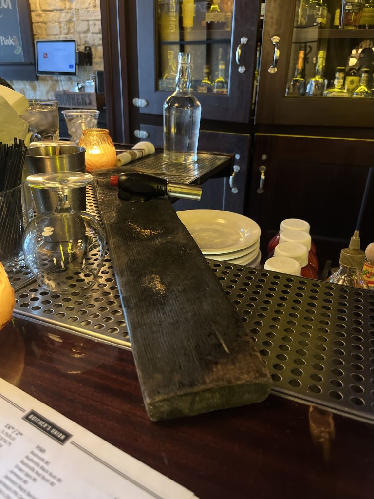 A charred board across the corner of a bar, used for adding smoke to cocktails.