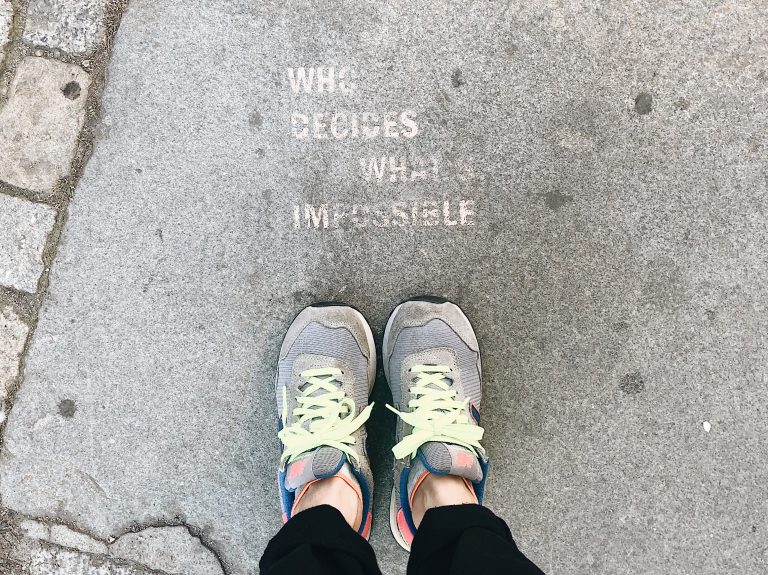 Person wearing running shoes in front of the text “who decides what’s impossible” made with a spray technique on the sidewalk