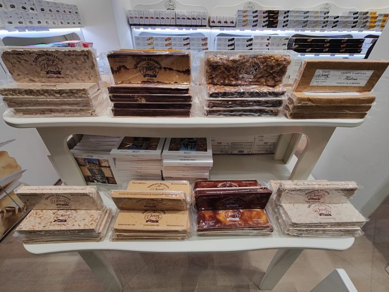 A variety of traditional Spanish turrones (nougats) displayed on a shelf, with flavors like almond, chocolate, and creamy nougat, all packaged and ready for sale.