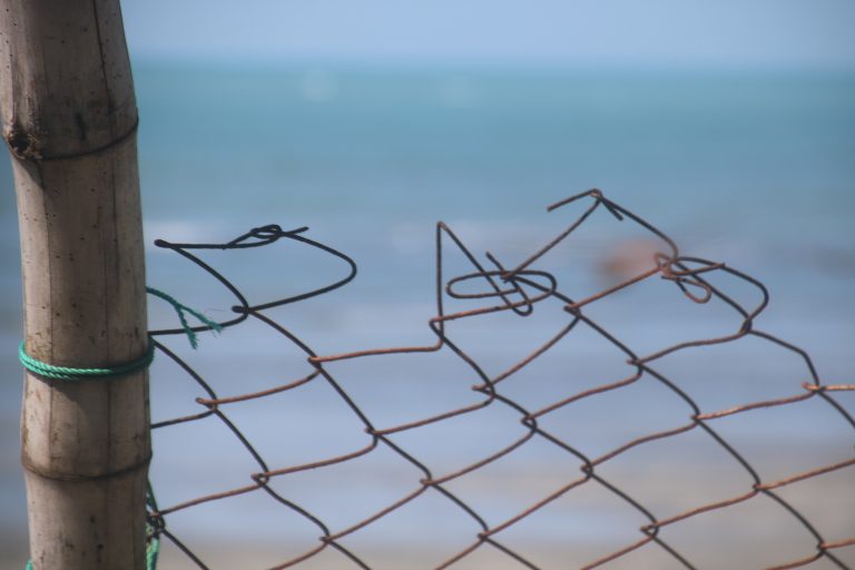 Ragged top of chainlink fence connected to a bamboo pole.  Ocean blurry in the background.