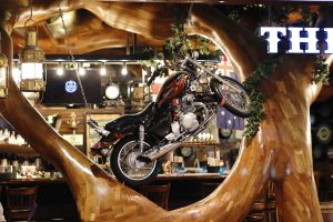 A black motorcycle with flame designs is mounted on a polished wooden structure resembling a tree in a bar setting, with an Australian flag, pendant lights, and shelves of bottles in the background

