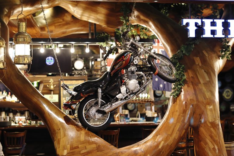 A black motorcycle with flame designs is mounted on a polished wooden structure resembling a tree in a bar setting, with an Australian flag, pendant lights, and shelves of bottles in the background