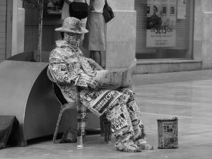 Man sitting in a chair on the street completely covered in newspaper, even his face.
