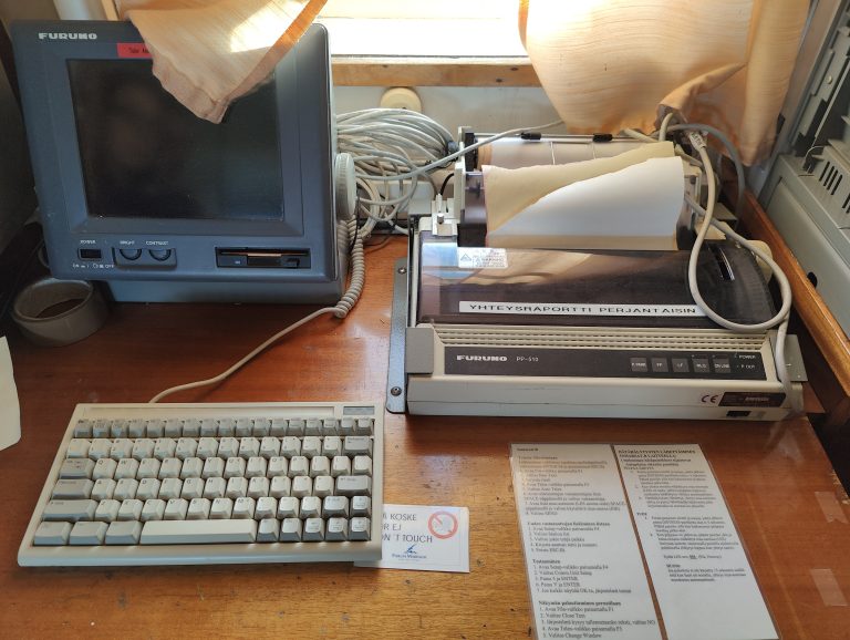 A vintage ship communication setup with a CRT monitor, keyboard, and printer, along with printed instructions, likely for maritime navigation and communication.