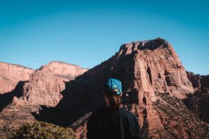 Person with a hat on, looking out at Zion National Park and the red colored mountain cliffs. 
