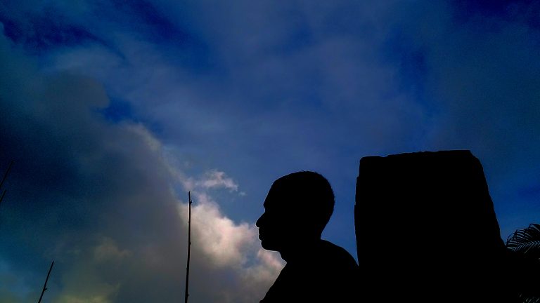 A boy is seated next to a pillar under the dark blue sky.