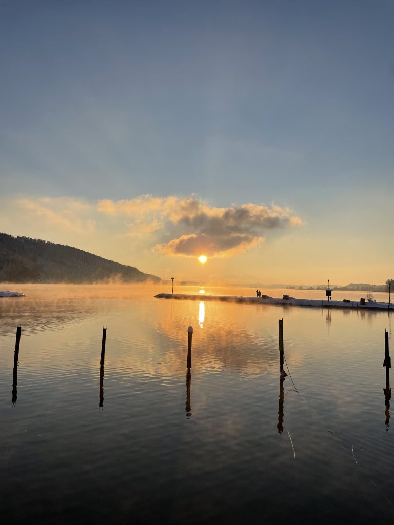 Sunset over a calm lake with silhouettes of people in the distance, wooden poles sticking out of the water, and mist rising from the surface against a backdrop of mountains and a sky with scattered clouds.