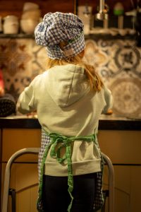 A young girl helping in the kitchen, standing on kitchen stairs, wearing a chef's hat and apron, seen from the back.