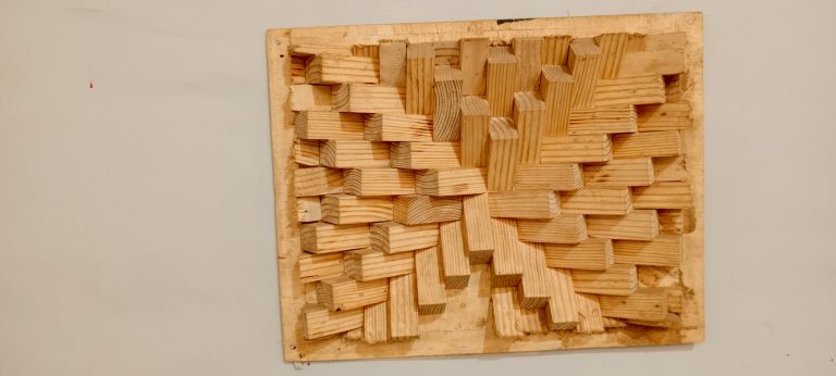 This photo displays a geometric wooden block sculpture against a white wall. It was taken at Motiv Uganda, Kampala.