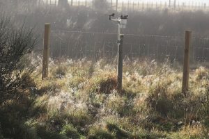 Weather station on a wire fence, dewdrops cling to the metal runners and cobweb laiden with dew glisten in the grass below. Taken on misty morning as the sun was breaking through the mist.