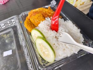 A plastic container with rice, cucumber, chicken, and a spoon. A delicious and refreshing meal ready to be enjoyed!