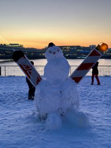 A life size snowman which has traffic delineators as arms and the Rhine river in the background at sunset.