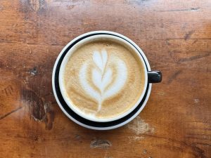 An overhead view of a cappuccino in a mug with milk art on a worn, dark wooden counter. 