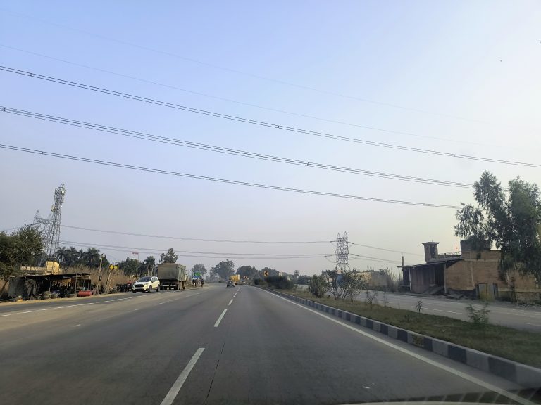 NH 19 highway view: vehicles moving along, power lines above, and scattered shops dotting the roadside, creating a vibrant urban scene.