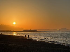 Sandy beach of Rethymno, Crete with people walking close to the water during sunset. In the background you can see the outlines of the old town and the Fortezza in front of some mountains.