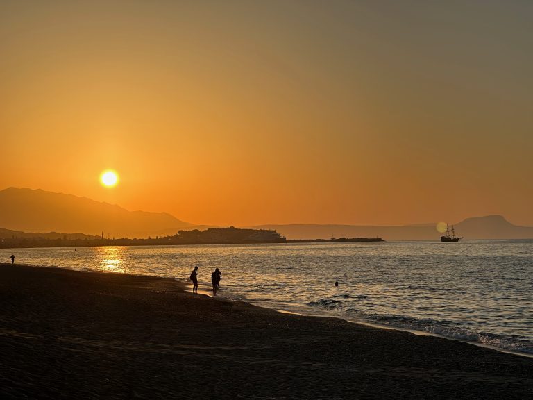 Sandy beach of Rethymno, Crete with people walking close to the water during sunset. In the background you can see the outlines of the old town and the Fortezza in front of some mountains.