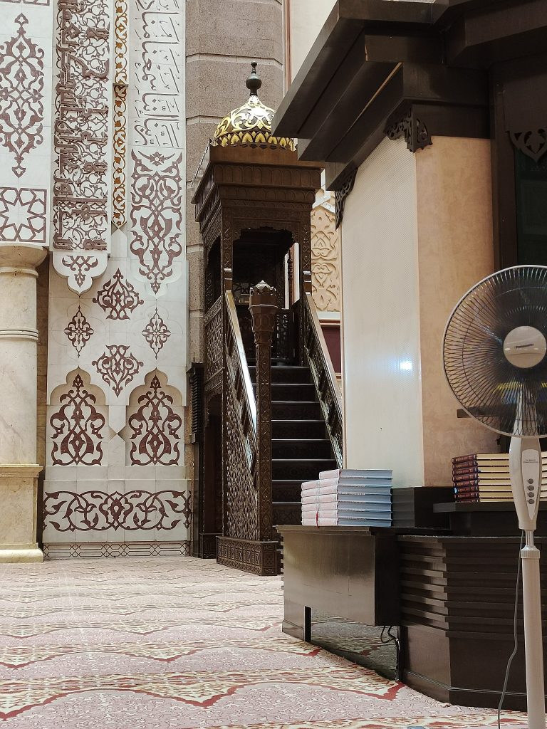 The wall with art works inside a mosque with a fan and a few books.