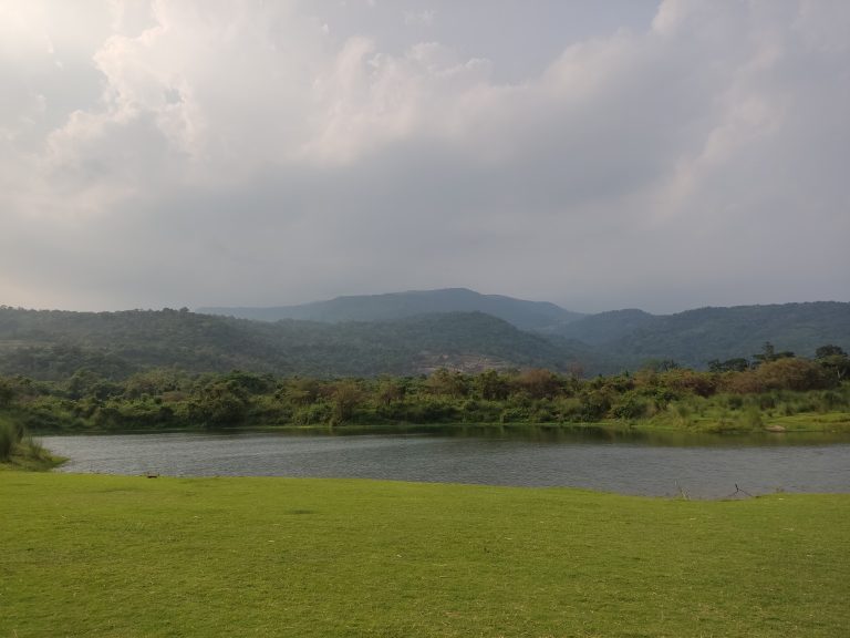 A  green field and lake in front of multiple mountains with a cloudy sky