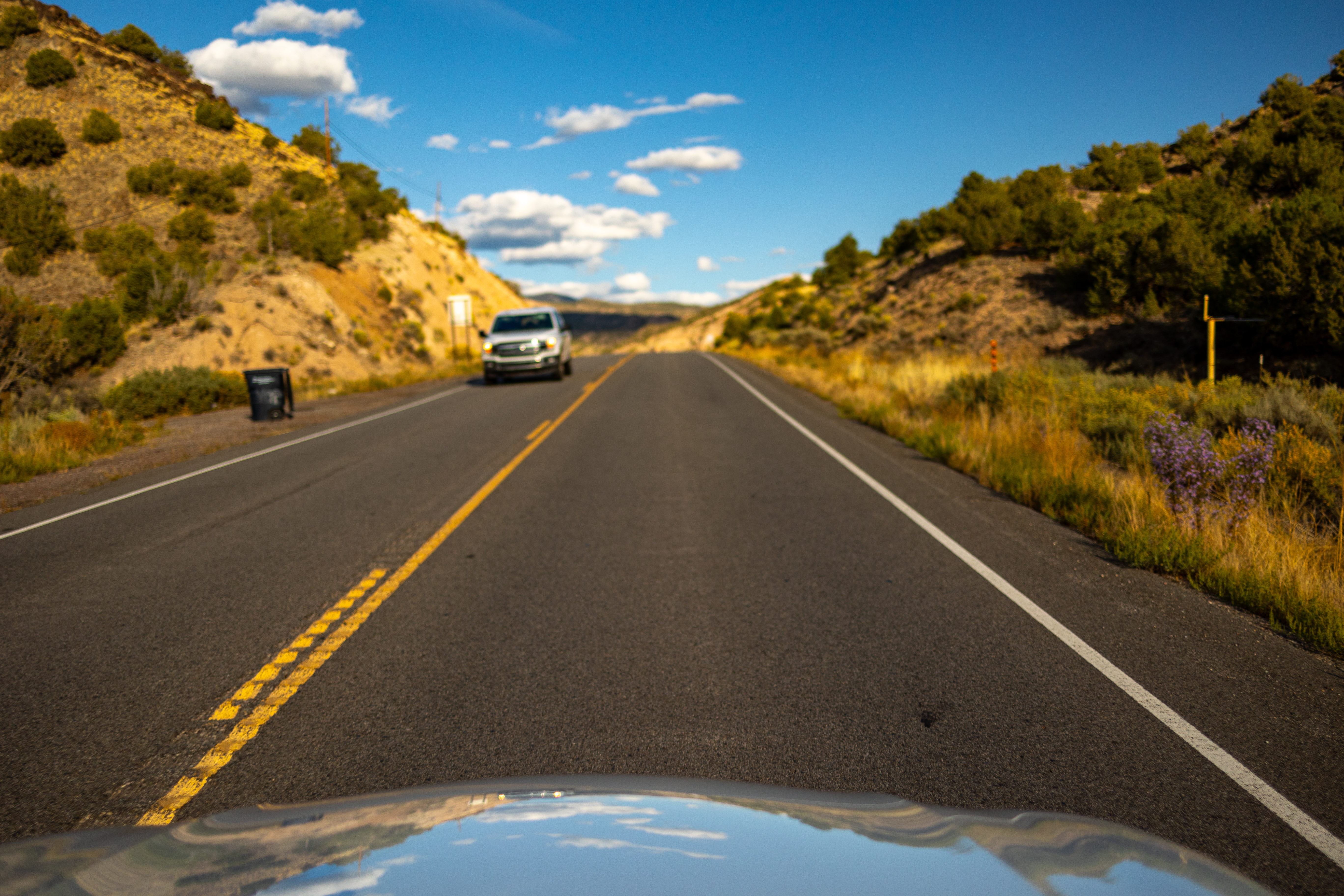 The view out of a car driving down a two lane road in a hilly high desert area of New Mexico with a truck approaching and a very blue sky.