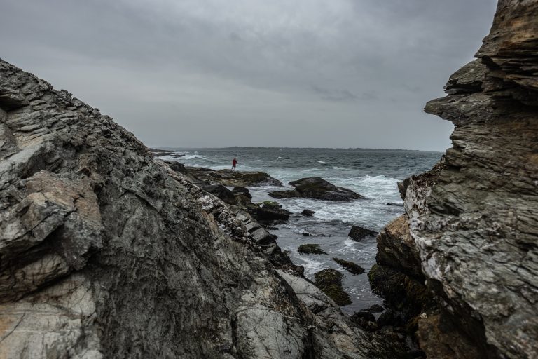 Looking through a rock formation at a seaside fisherman courageously casts his line as a storm approaches Beavertail Lighthouse in Rhode Island.