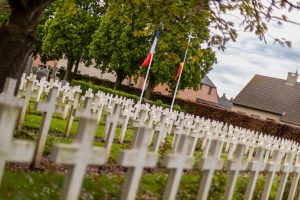 Rows of crosses in a World War 1 cemetery in Belgium with French and Belgian flags. Trees and houses are in the background. 
