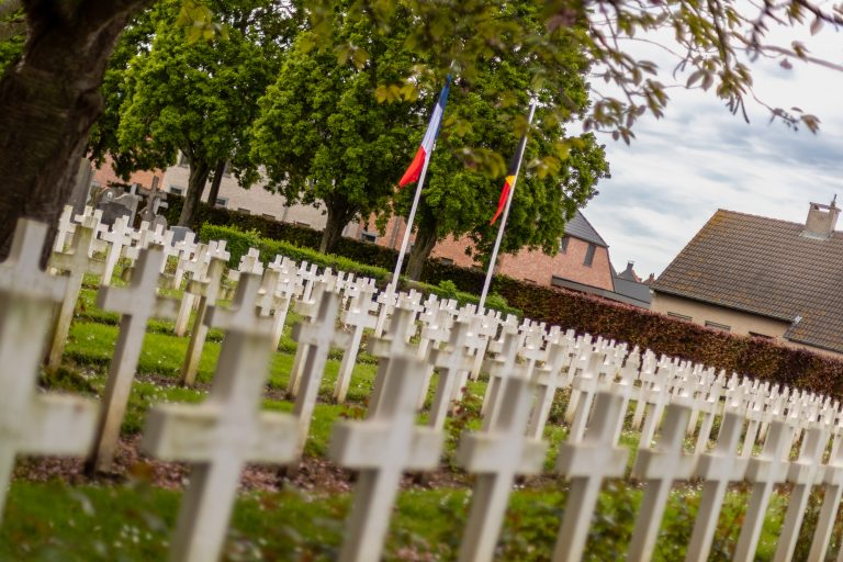 Rows of crosses in a World War 1 cemetery in Belgium with French and Belgian flags. Trees and houses are in the background.