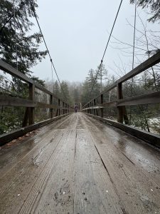 In Lincoln, New Hampshire, a woman in a purple winter jacket stands on a wooden suspension bridge over the East Branch Pemigewasset River in the rain.