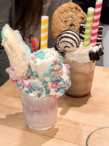 Two gluttonous ice cream shakes. One is pink with cotton candy and a marshmallow on top. The other is chocolate with a full chocolate chip cookie and a cake pop on top. Both have rims covered in whipped cream with colorful chocolate pieces and wide straws sticking out.
