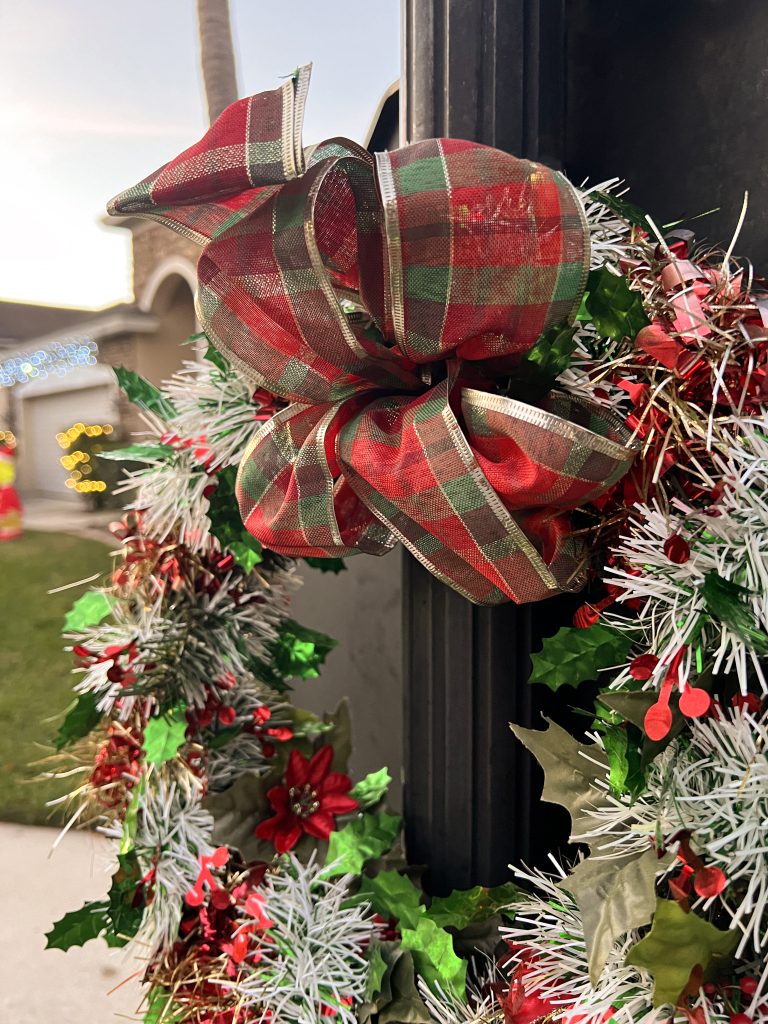 Close shot of a white tinsel Christmas wreath with fake red flowers and green leaves. It also has a red and green plaid bow and is hanging on a street sign pole.