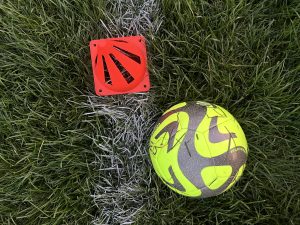 A neon green soccer ball and a neon orange cone rest on a line against a dark green grass background.