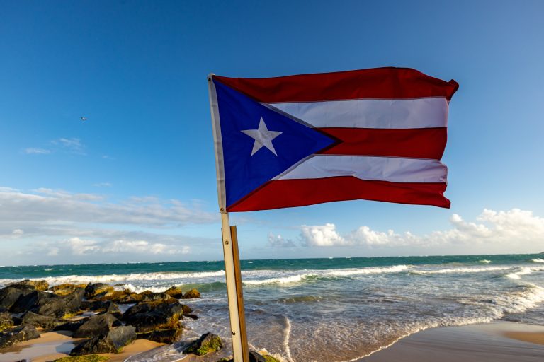 Puerto rican flag blowing in the wind on a beach with with ocean in the background and a small outcropping of rocks on the left side