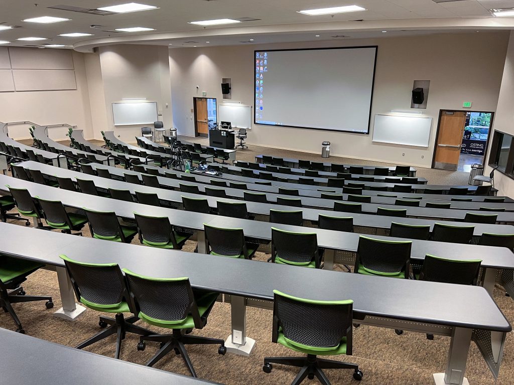 Empty college classroom with 10 rows of chairs on layered levels. Podium and projector showing computer desktop at the front of the class.