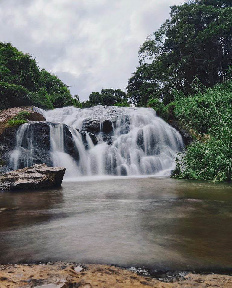 The river cascades down rocky cliffs, surrounded by towering trees and vibrant green foliage, and the sky above filled with clouds. Kotagiri Waterfall, Tamil Nadu.