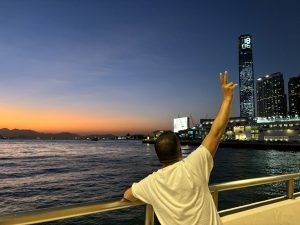 A person showing the peace sign standing on a bridge above a river at evening, in the distance you can see the city lights