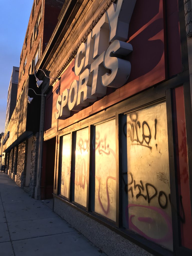 An abandoned City Sports store front on Commonwealth Avenue in North Brookline, MA. The windows are covered on the inside and there’s graffiti on the outside. The sun is setting and shining warm light on the building.