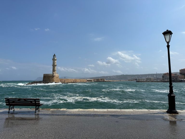 The Venetian Harbor of Chania city with the old sea wall and the lighthouse as viewed from the promenade between a bench and a streetlight.