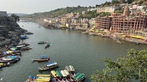 The image shows a large body of water, most likely the narmada river in india, with a ghat and several boats on it. The boats are mostly small traditional wooden boats, painted in bright colors. There are also a few larger boats, including a houseboat. 
