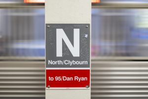 Scratched sign for the Chicago El Red Line to 95/Dan Ryan at North/Clybourne on a white pillar with a train moving in the background. 
