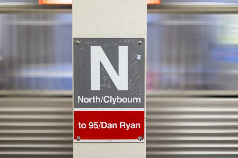 Scratched sign for the Chicago El Red Line to 95/Dan Ryan at North/Clybourne on a white pillar with a train moving in the background.