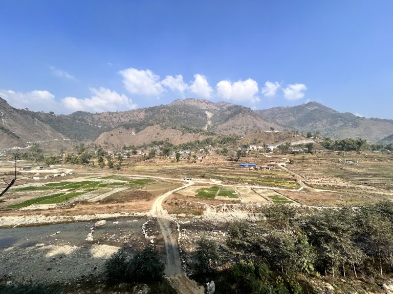 A glimpse of an uneven riverside road, surrounded by lush green fields, villages, and a clear sky with scattered clouds.